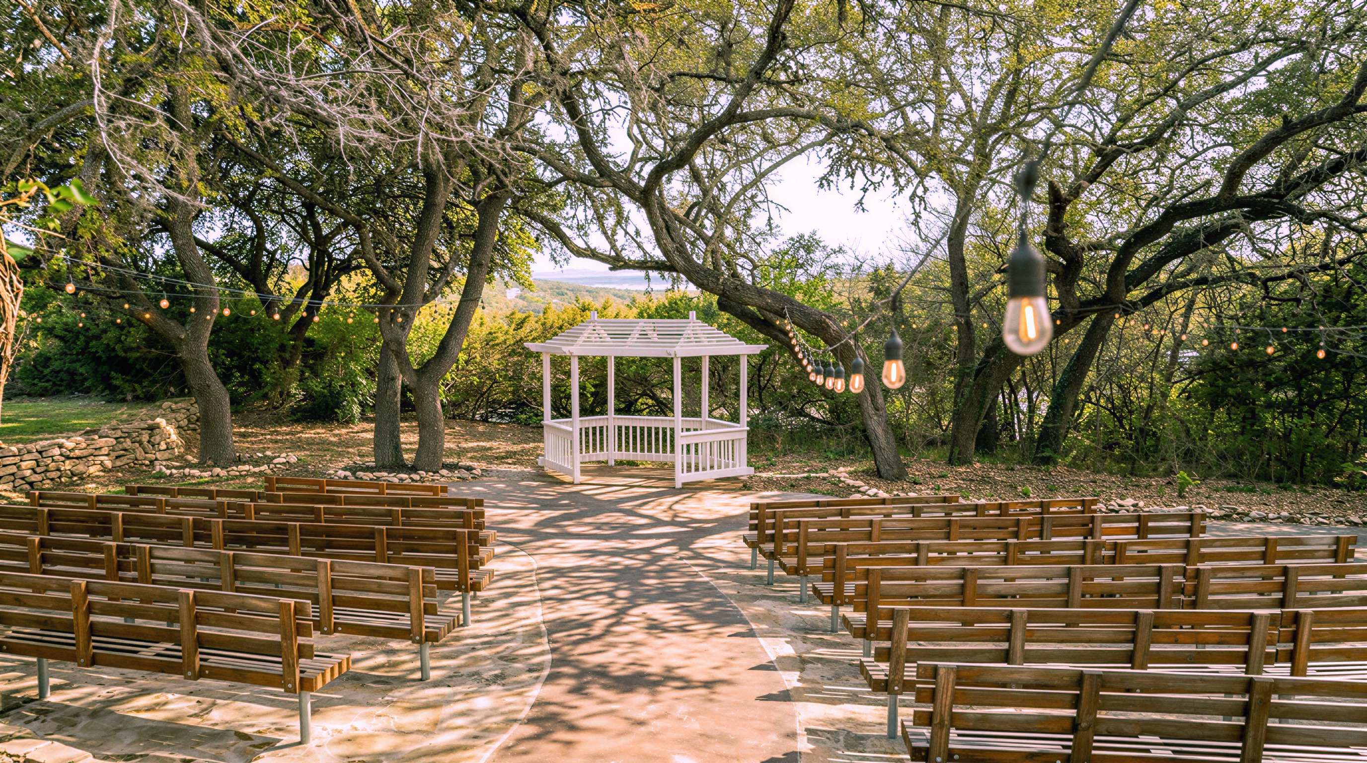 Rows of benches curving toward the white gazebo under the oaks, string lights overhead